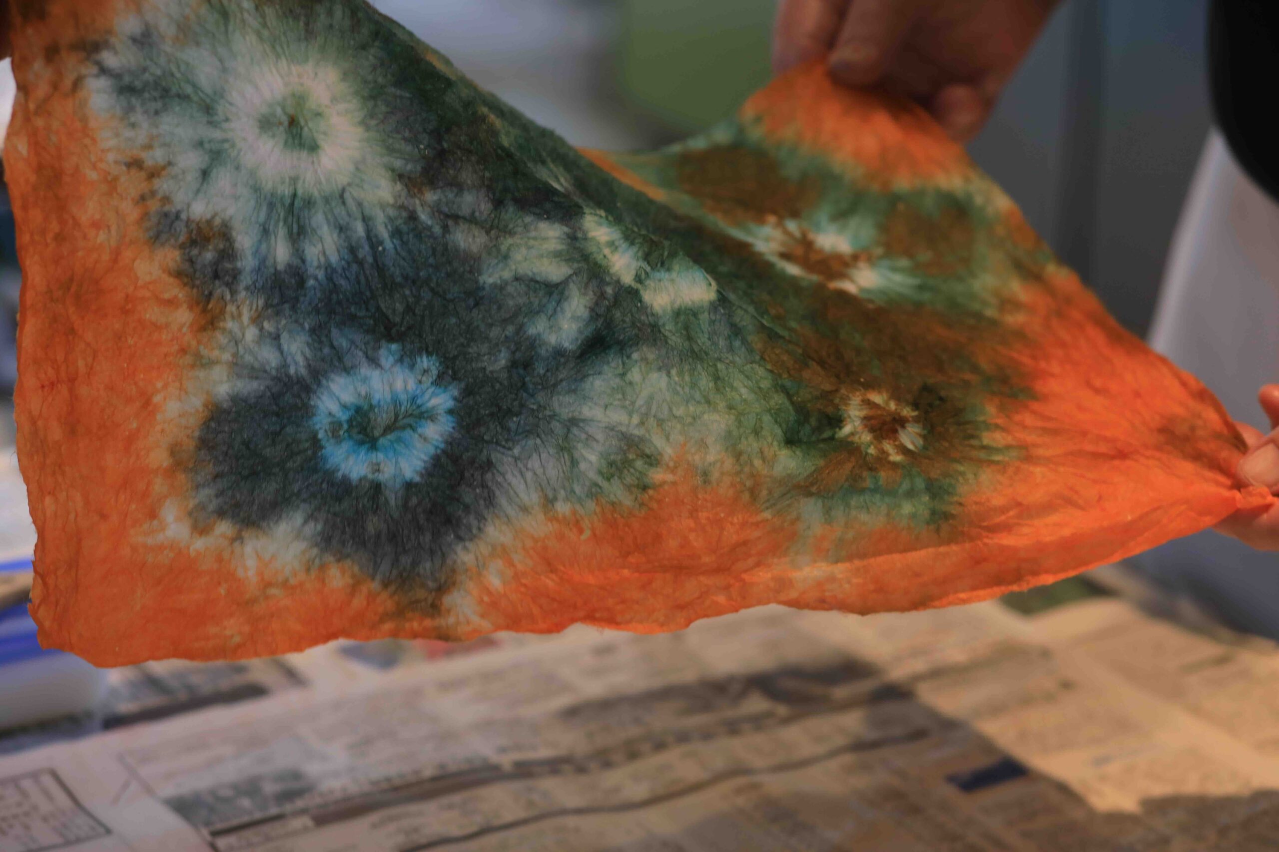 Finished handmade uchiwa fan with a bold orange, teal, and purple tie-dye pattern, held up at the end of the fan-making workshop